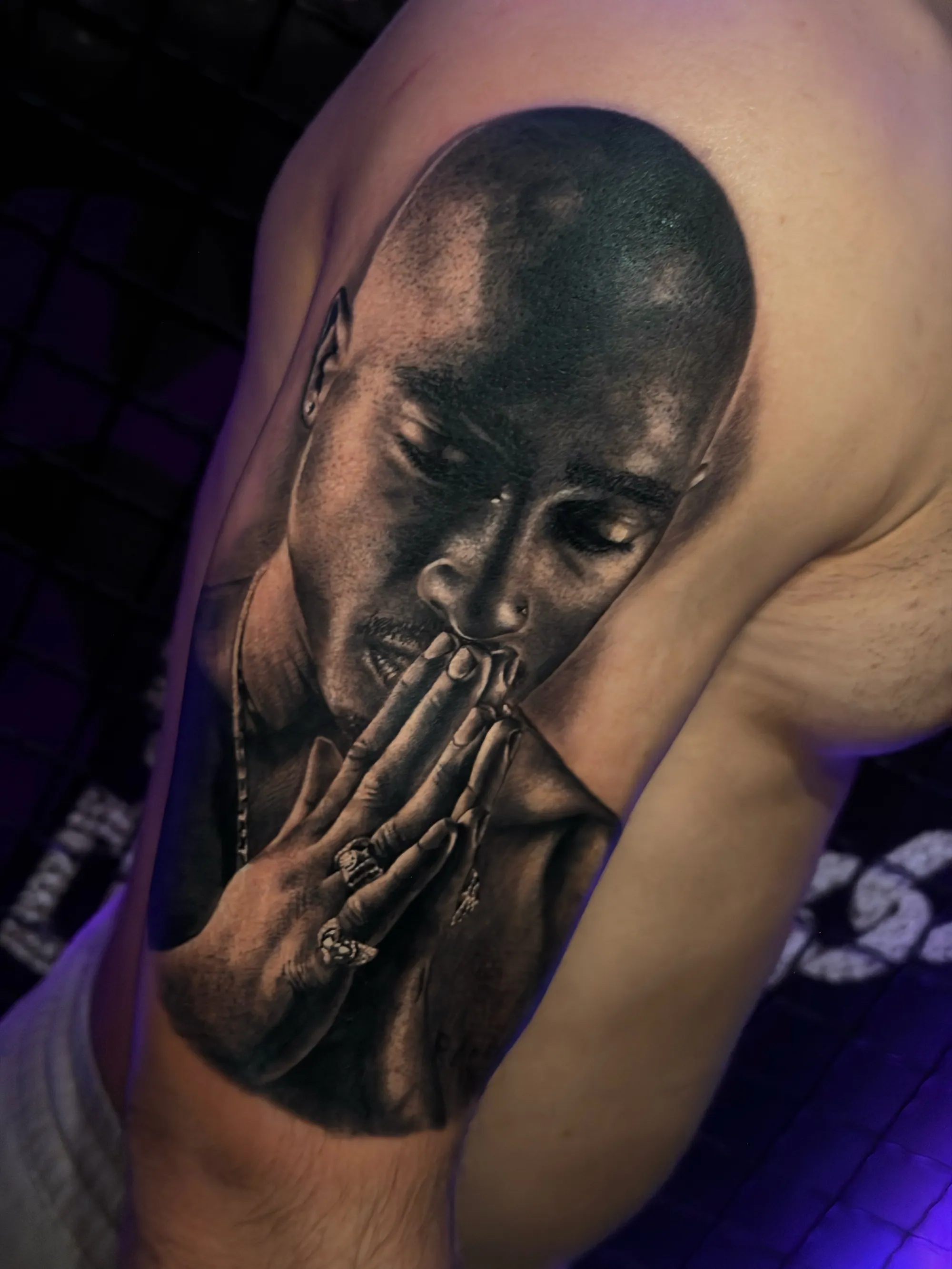 Realistična crno-bela tetovaža portreta Tupaca Shakura sklopljenih ruku na ramenu, Dropz Tattoo Studio Niš
